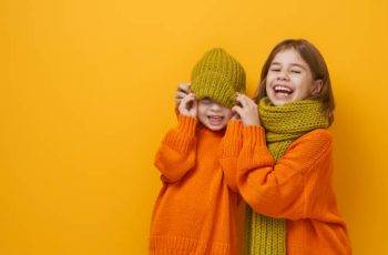 Winter portrait of happy children wearing knitted hat, snood and sweaters. Girls having fun, playing and laughing on yellow background. Fashion concept.